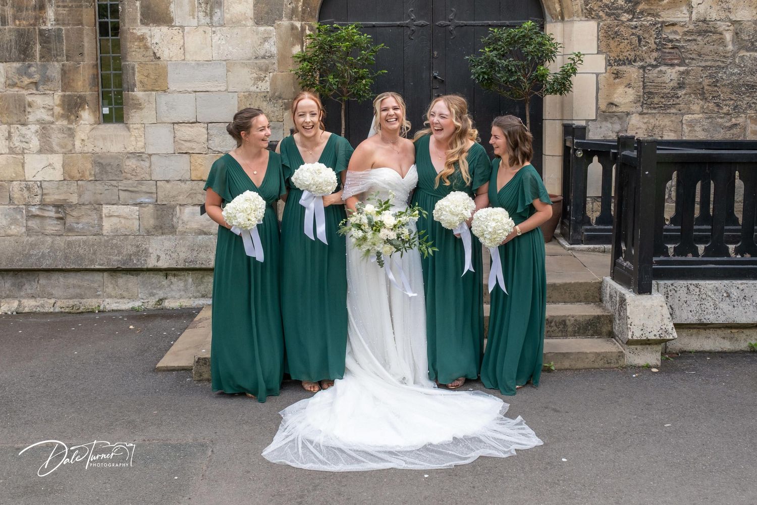 Wedding group photo session outside historic stone church with bridal party in green and white attire.