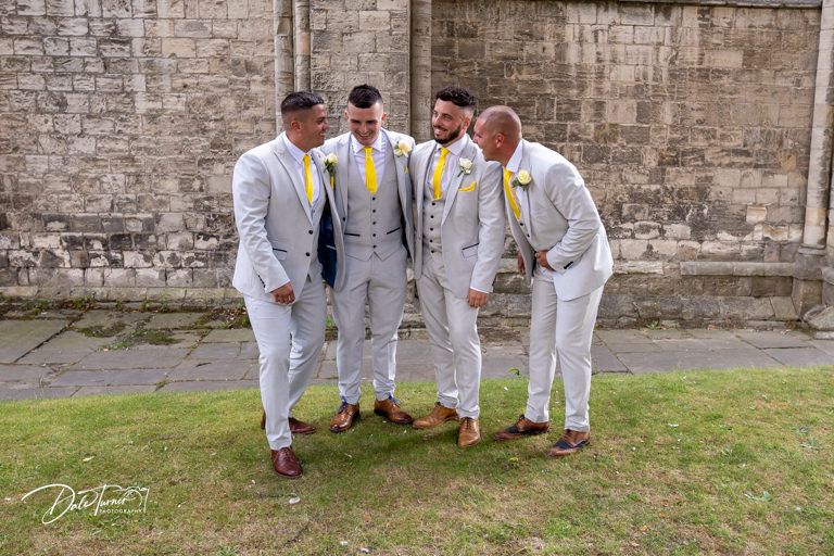 Groomsmen in white suits gather together against a stone wall backdrop at a wedding celebration.