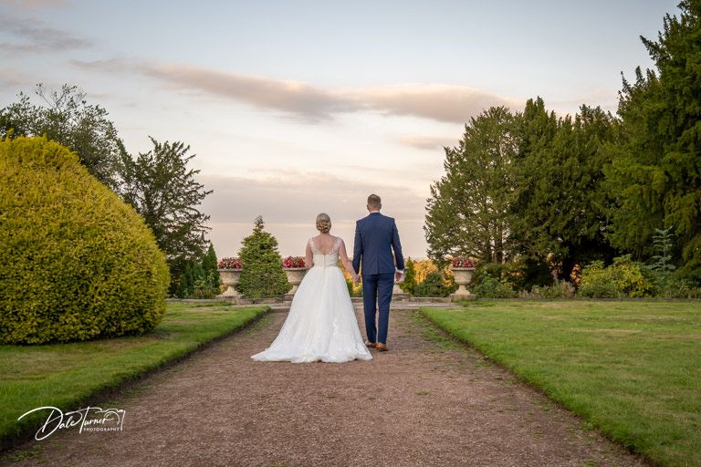 Wedding couple walks hand in hand along scenic garden path at sunset.
