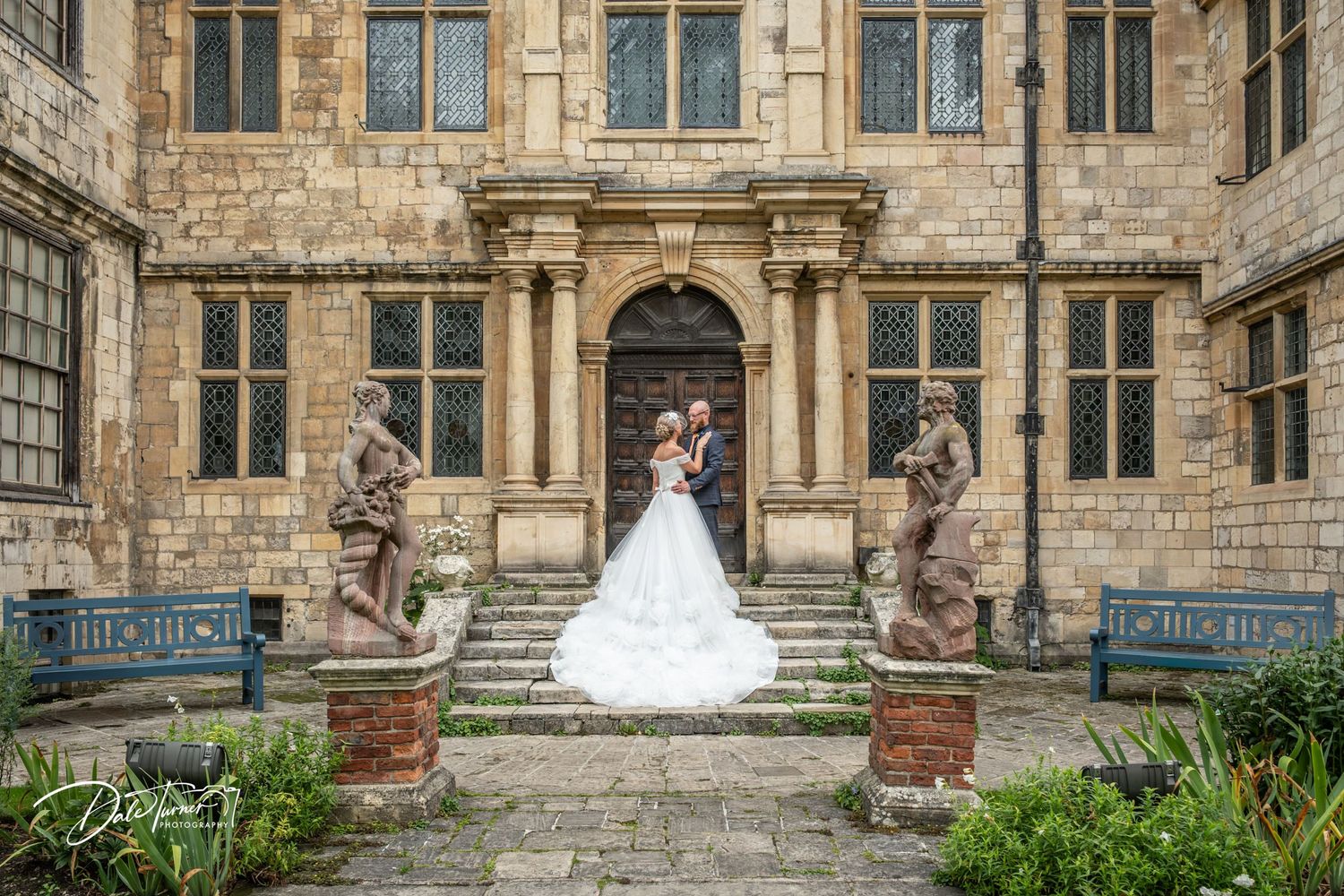 Bride and groom pose on stone steps of a grand historic manor, flanked by ornate stone sculptures in a courtyard.