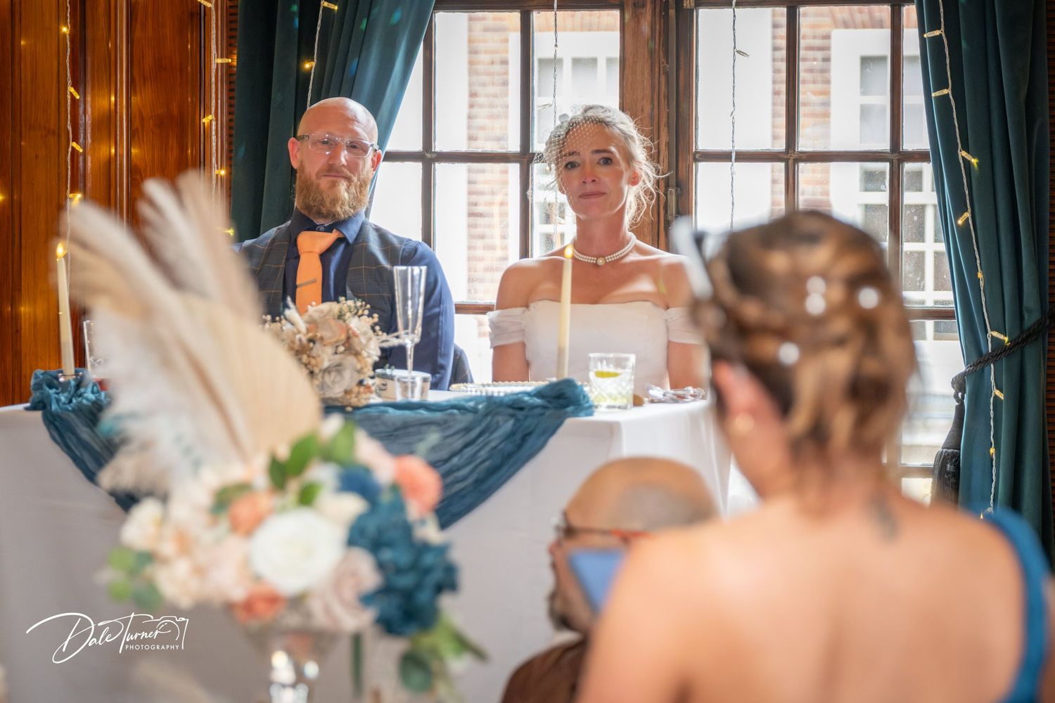 Bride and groom smiling at head table during elegant wedding reception with blue decor and fairy lights.