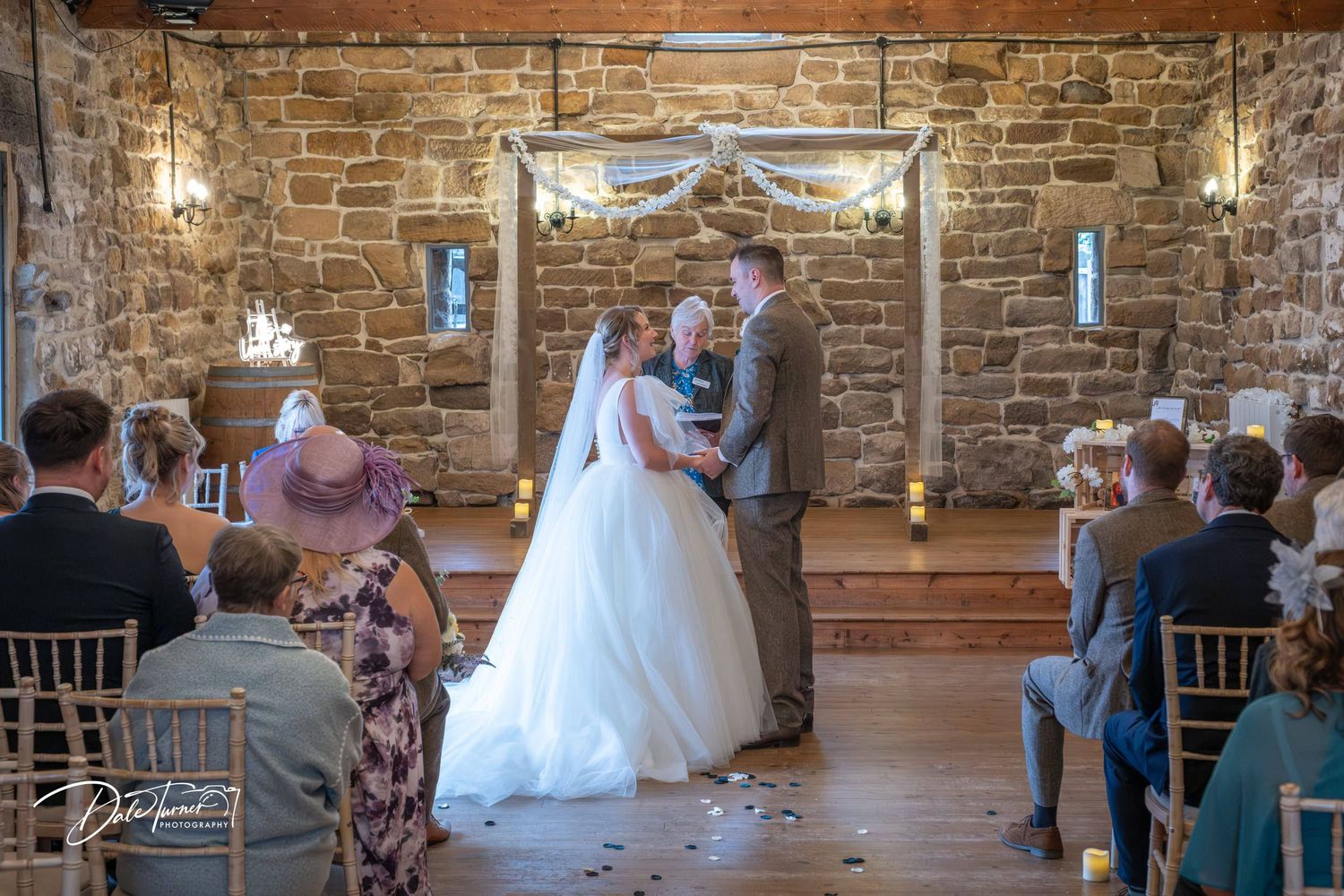 Wedding guests seated in a rustic stone venue with wooden beams and chandeliers during a ceremony.