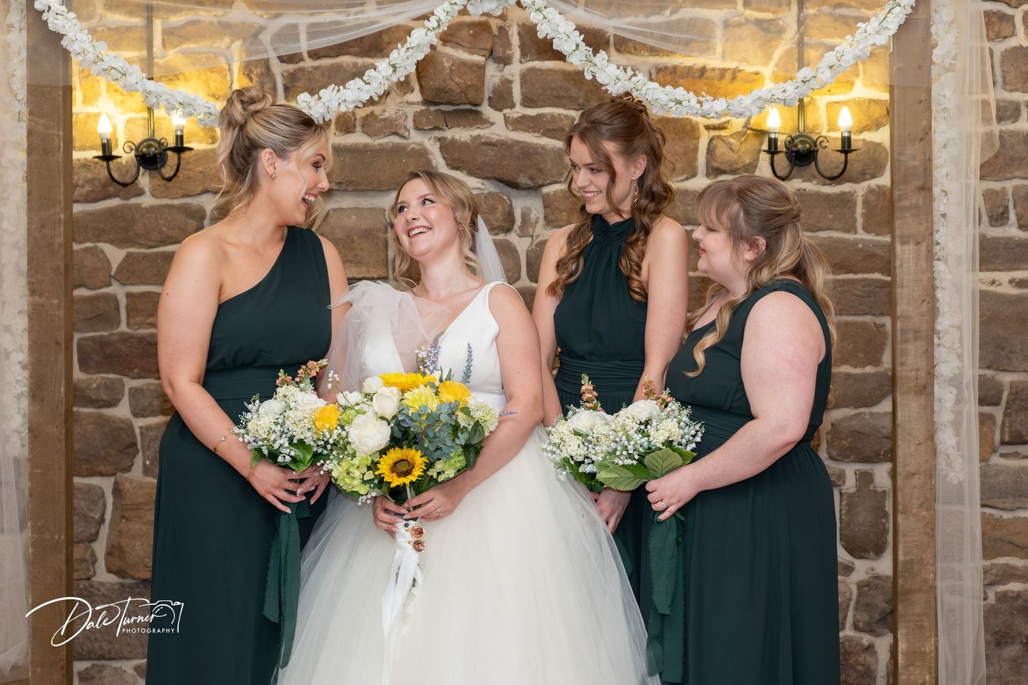 Wedding party in dark green dresses and white gown pose together in rustic brick venue with string lights overhead.