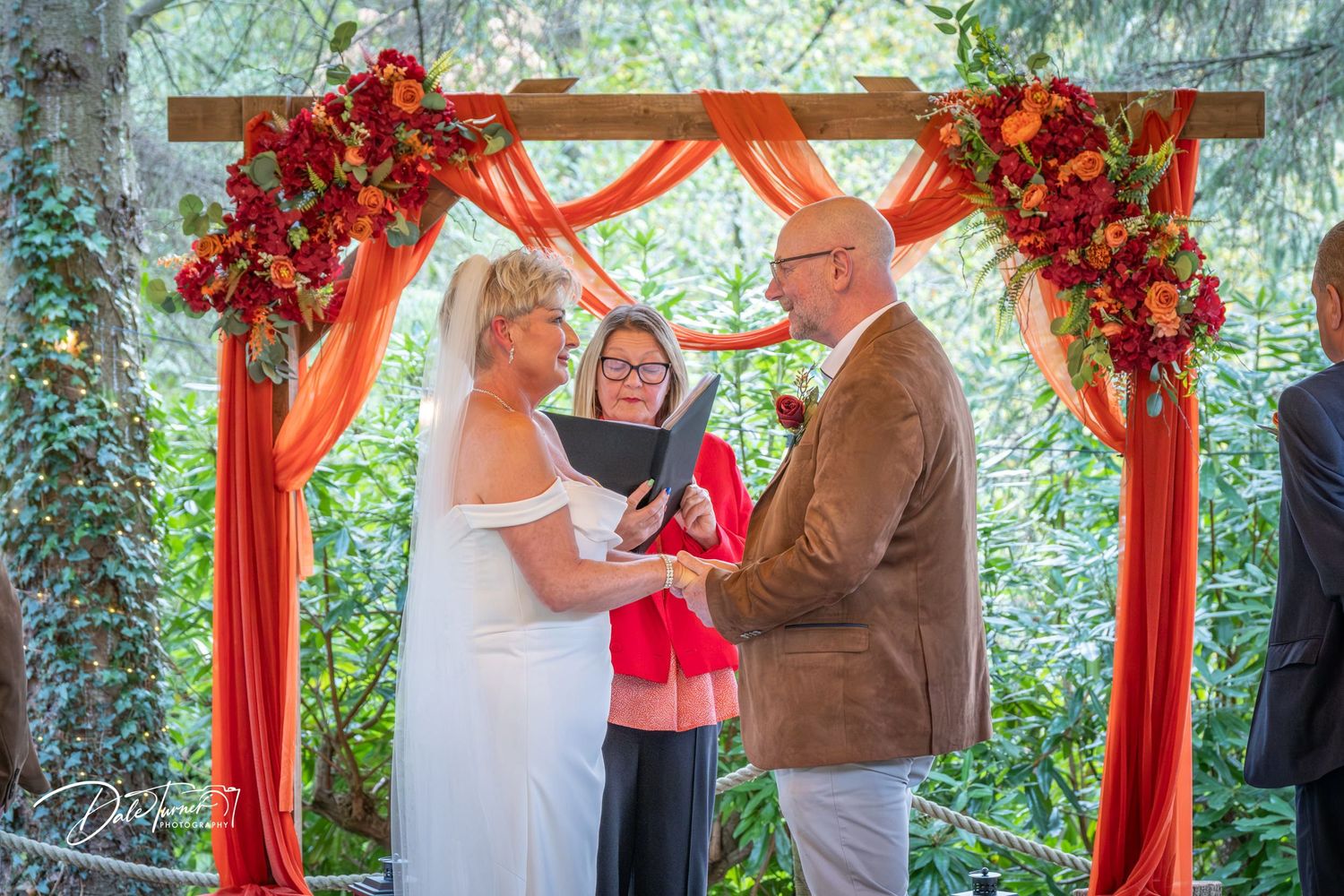 Intimate wedding ceremony taking place beneath a vibrant red floral archway.