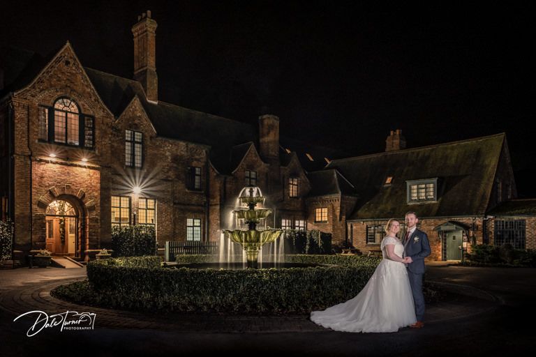 A romantic nighttime wedding portrait in front of an illuminated Victorian mansion.