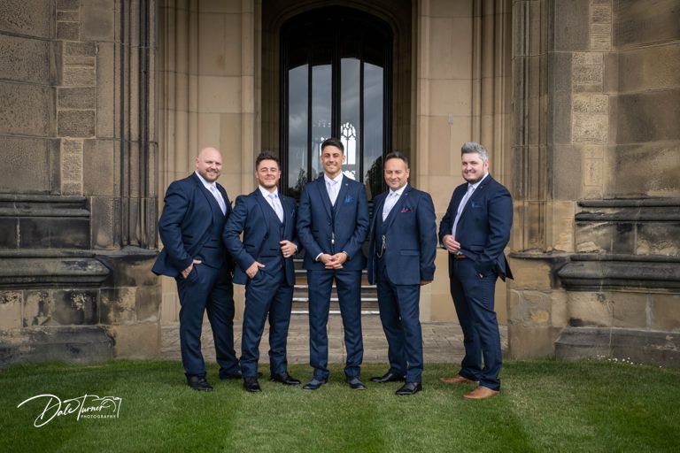 Wedding party in navy suits gathered outside historic stone architecture.