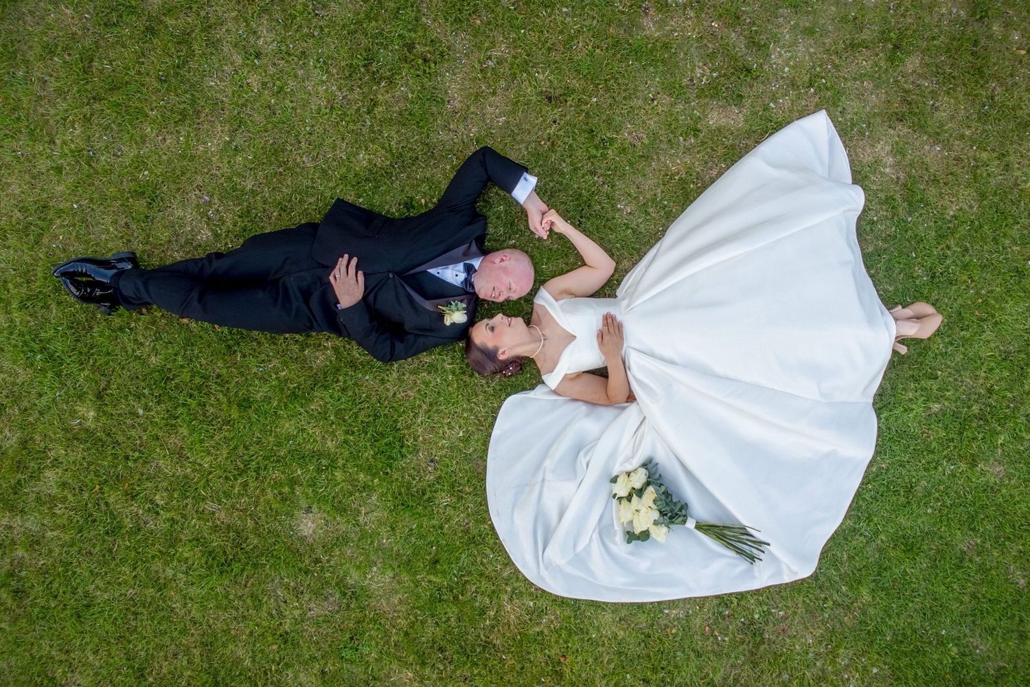 Overhead view of a couple in formal attire lying on green grass.