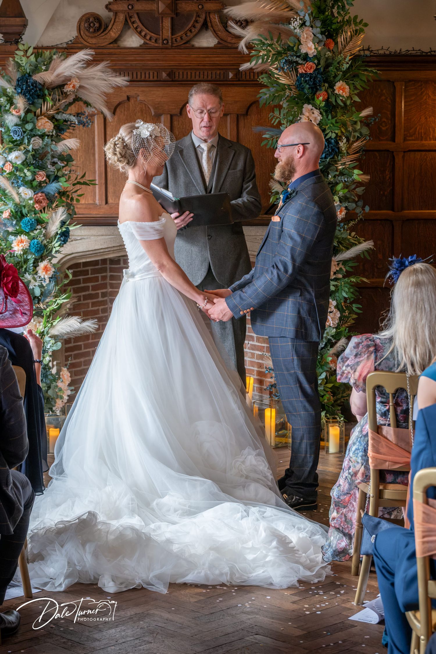 Bride and groom hold hands during indoor wedding ceremony before officiant, framed by floral arch with pampas grass.