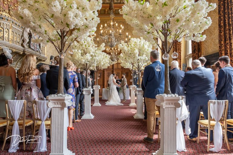 Wedding ceremony in historic hall with white cherry blossom trees lining the aisle.
