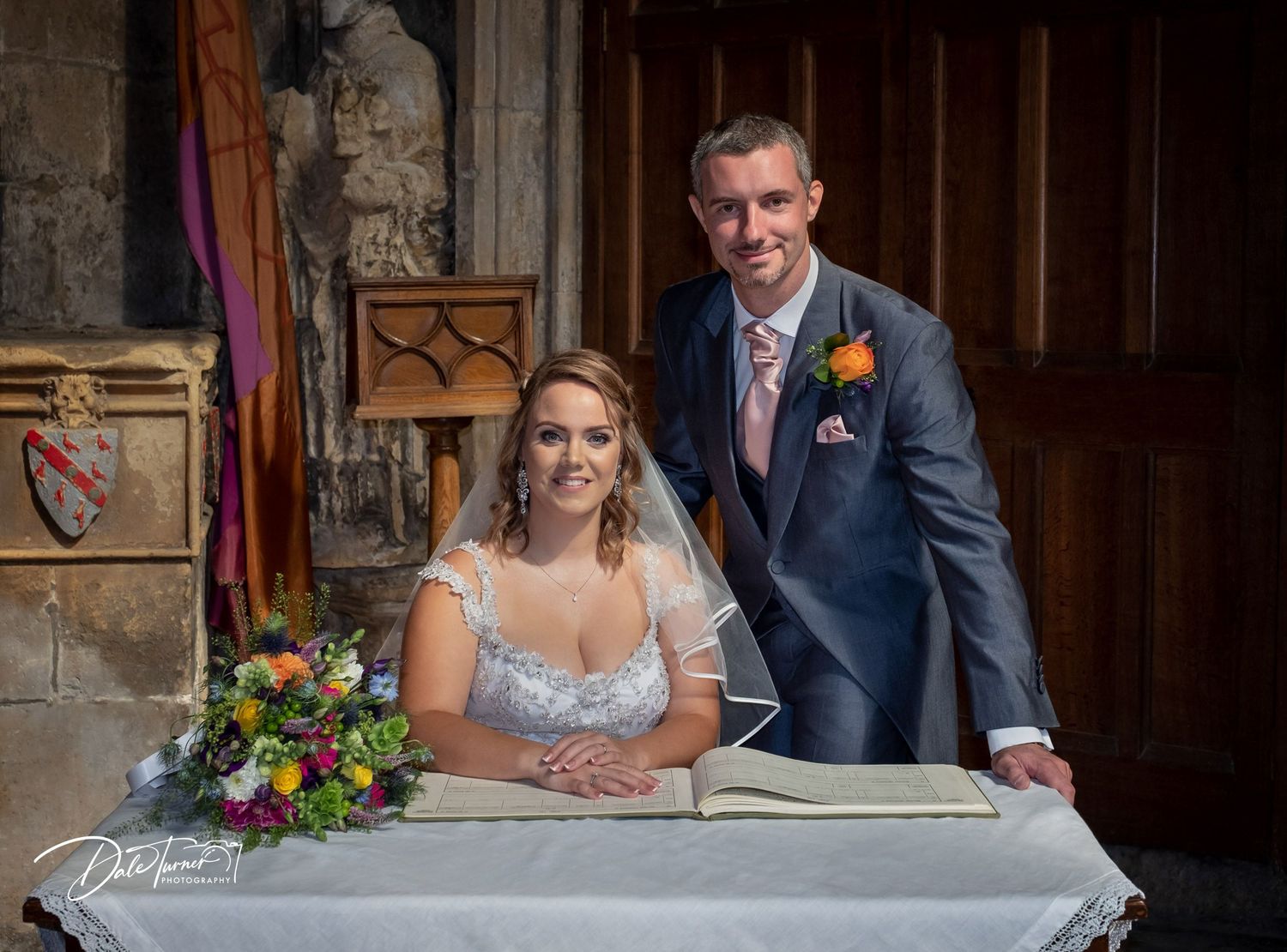Wedding couple signs documents at an ornate stone table in a historic building interior.