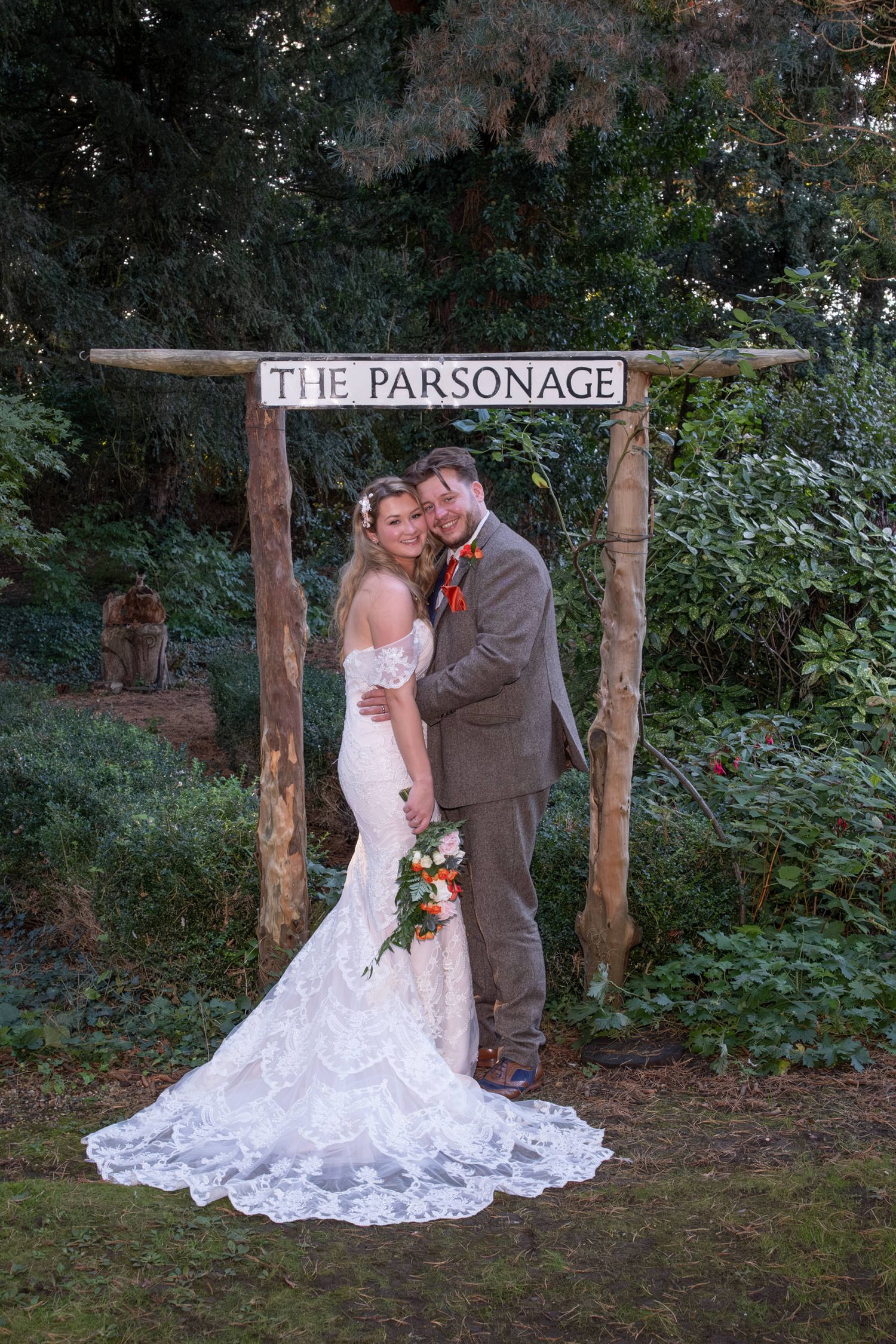 A romantic wedding photoshoot in front of a rustic wooden sign reading 'The Parsonage' surrounded by greenery.