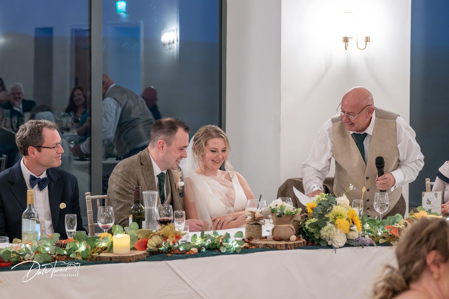 Wedding reception guests seated at long tables decorated with sunflowers and greenery during dinner service.