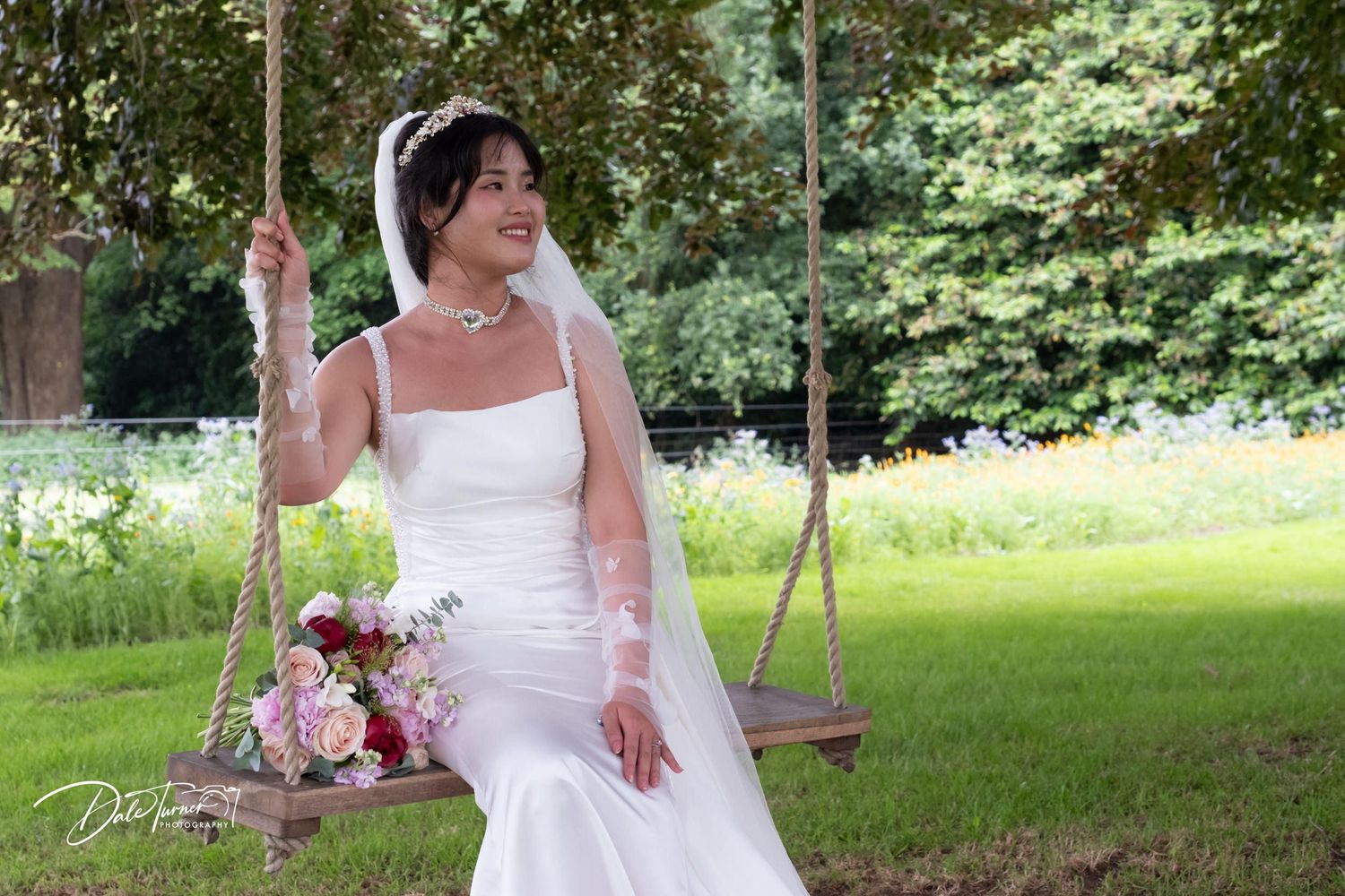 Bride in white dress sits on rustic wooden swing in lush garden setting.