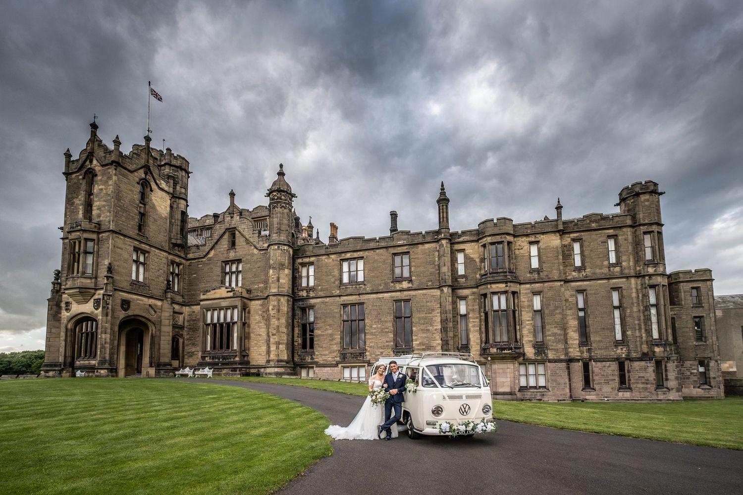 Dramatic panorama of Gothic castle architecture under stormy skies with classic car in foreground.