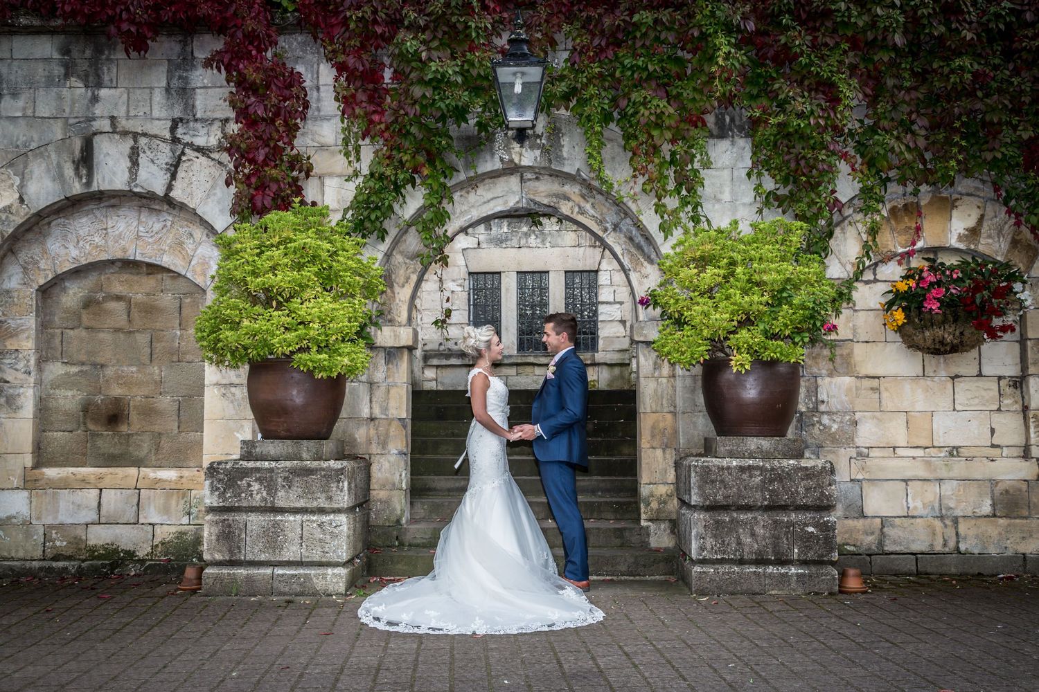 Wedding couple standing in front of ivy-covered stone archway with decorative planters.