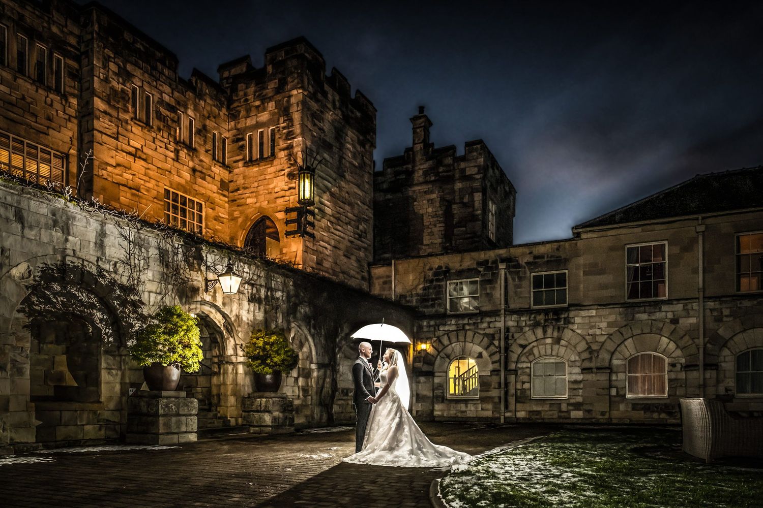 Dramatic nighttime wedding photo with white umbrella against illuminated stone castle walls.