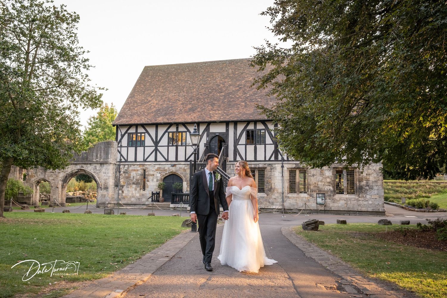 A couple poses in front of a historic Tudor-style building with stone archways.