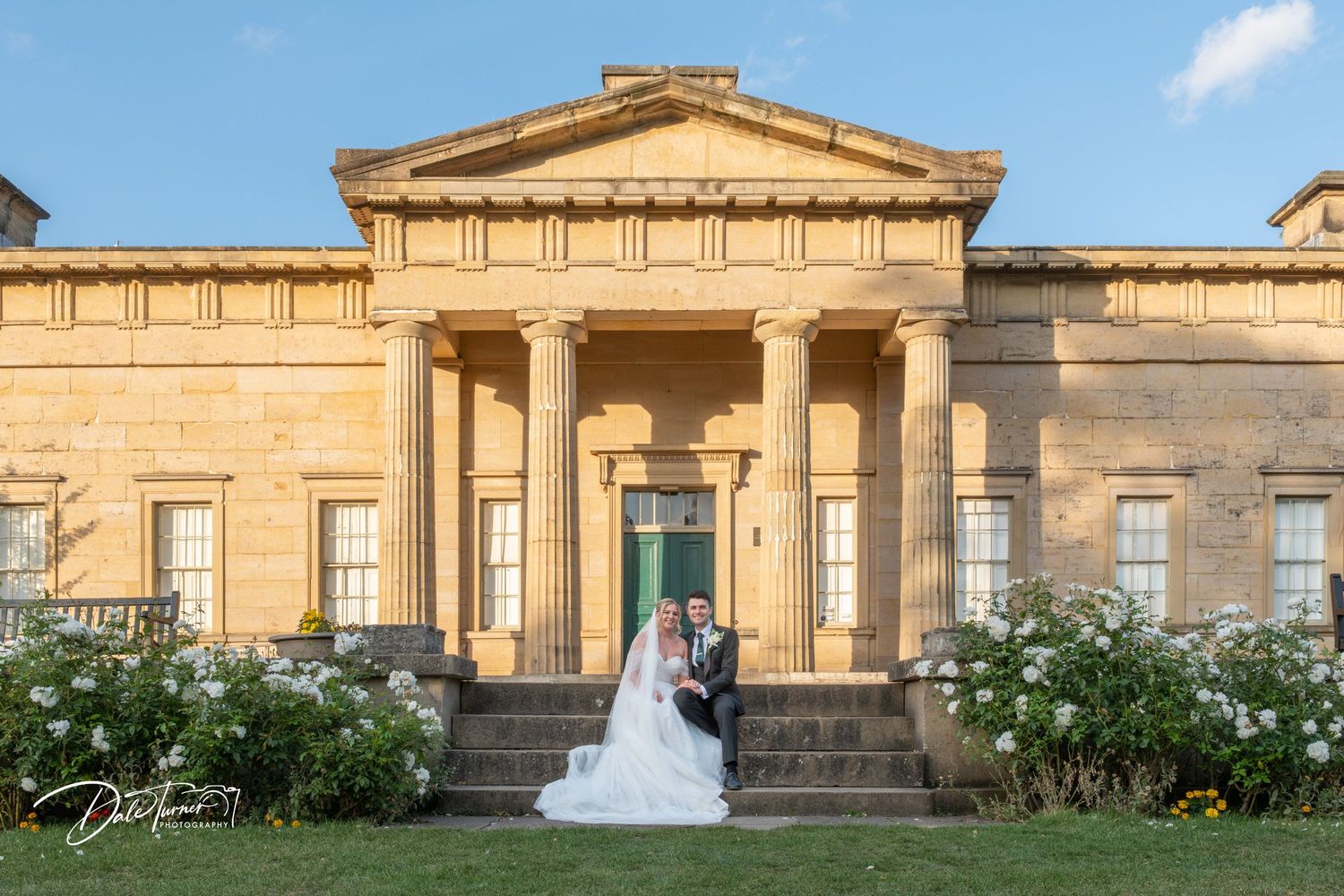 A wedding couple poses in front of a grand neoclassical building with columns and gardens.