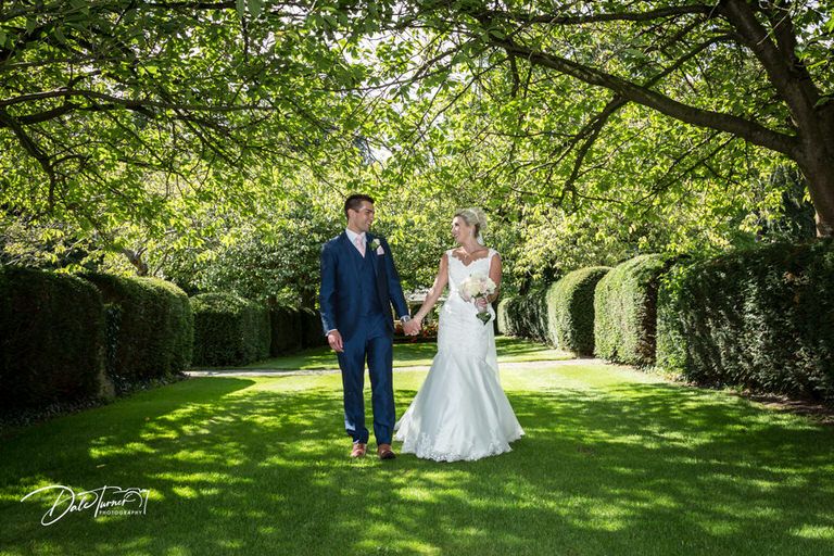 Wedding couple walks hand in hand through a lush green garden path lined with trees.