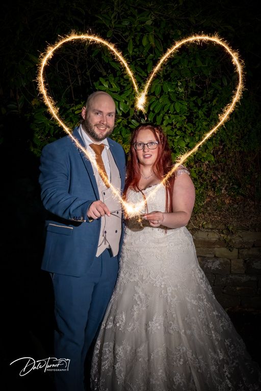 Wedding couple holding sparklers creating heart shapes against dark evening background.