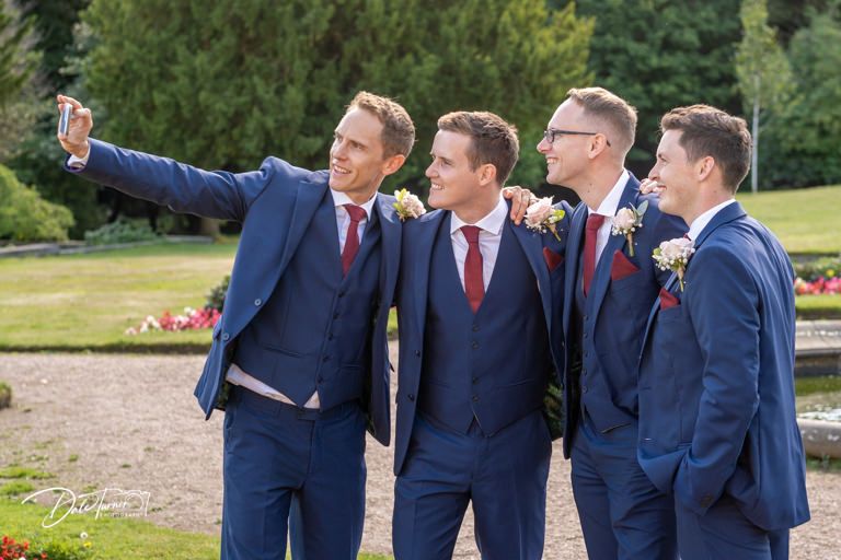 Groomsmen in navy suits taking a selfie together in a garden setting.