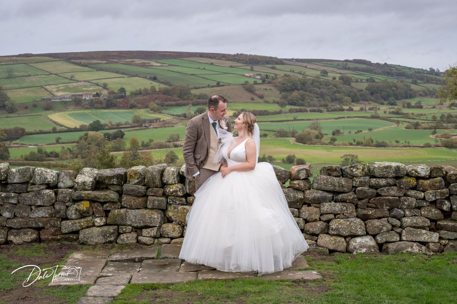 Wedding photography scene on a hilltop overlooking scenic green countryside with stone walls and rolling fields.