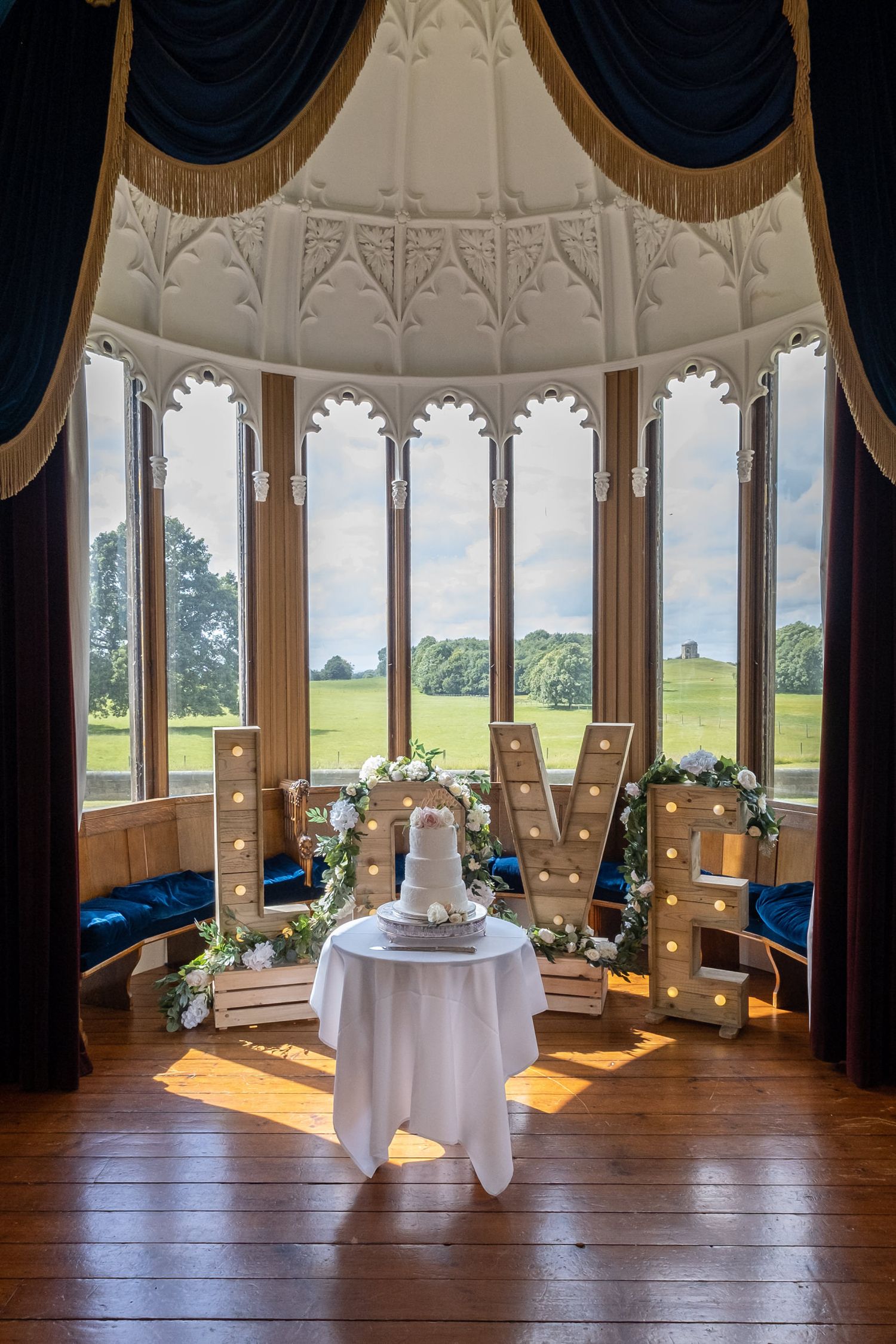 Elegant round wedding cake displayed in sunlit bay window room with dramatic drapery.
