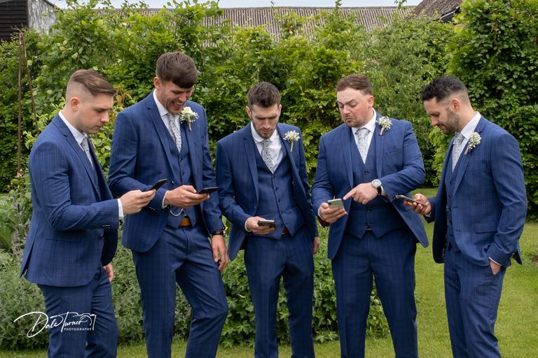 Groomsmen in navy suits check their phones in synchronized outdoor photo.