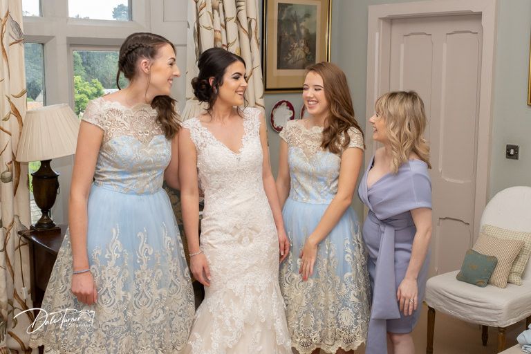 Bridesmaids in pastel blue and lavender dresses help with wedding preparations in an elegant room.