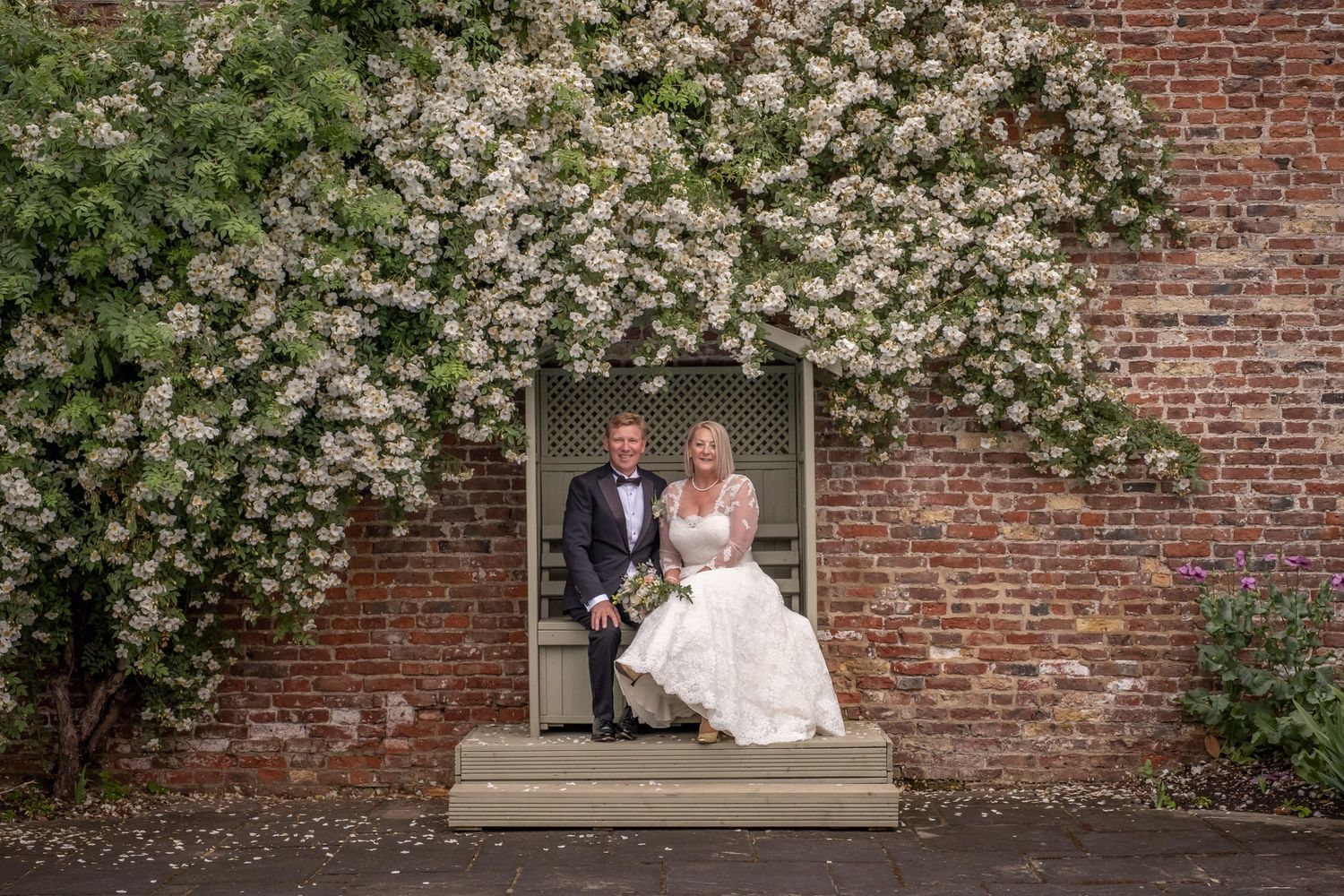 Wedding portraits against a classic brick wall adorned with blooming white flowers.