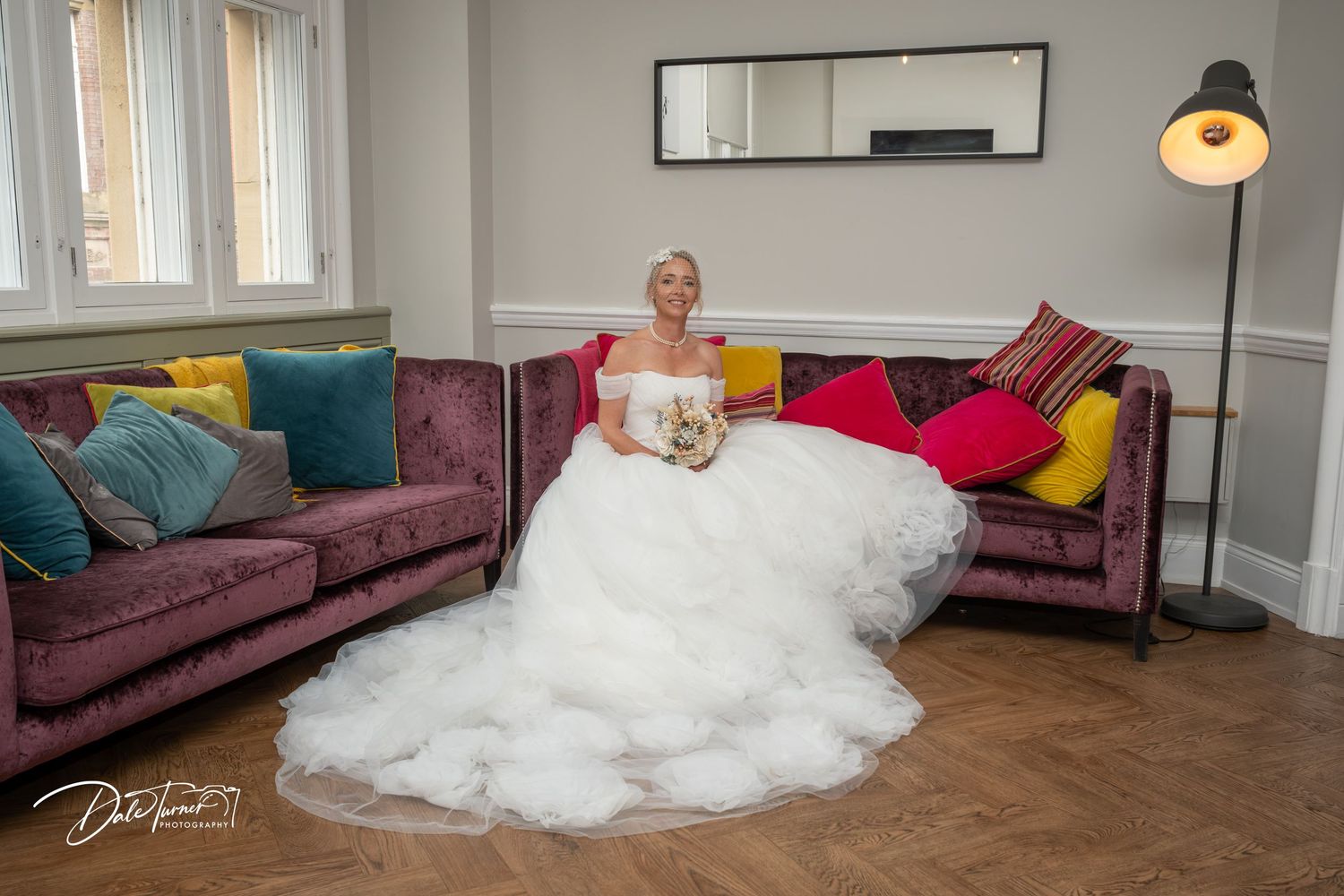 Bride in a white ballgown sitting on a purple velvet sofa with colorful cushions in an elegant room.