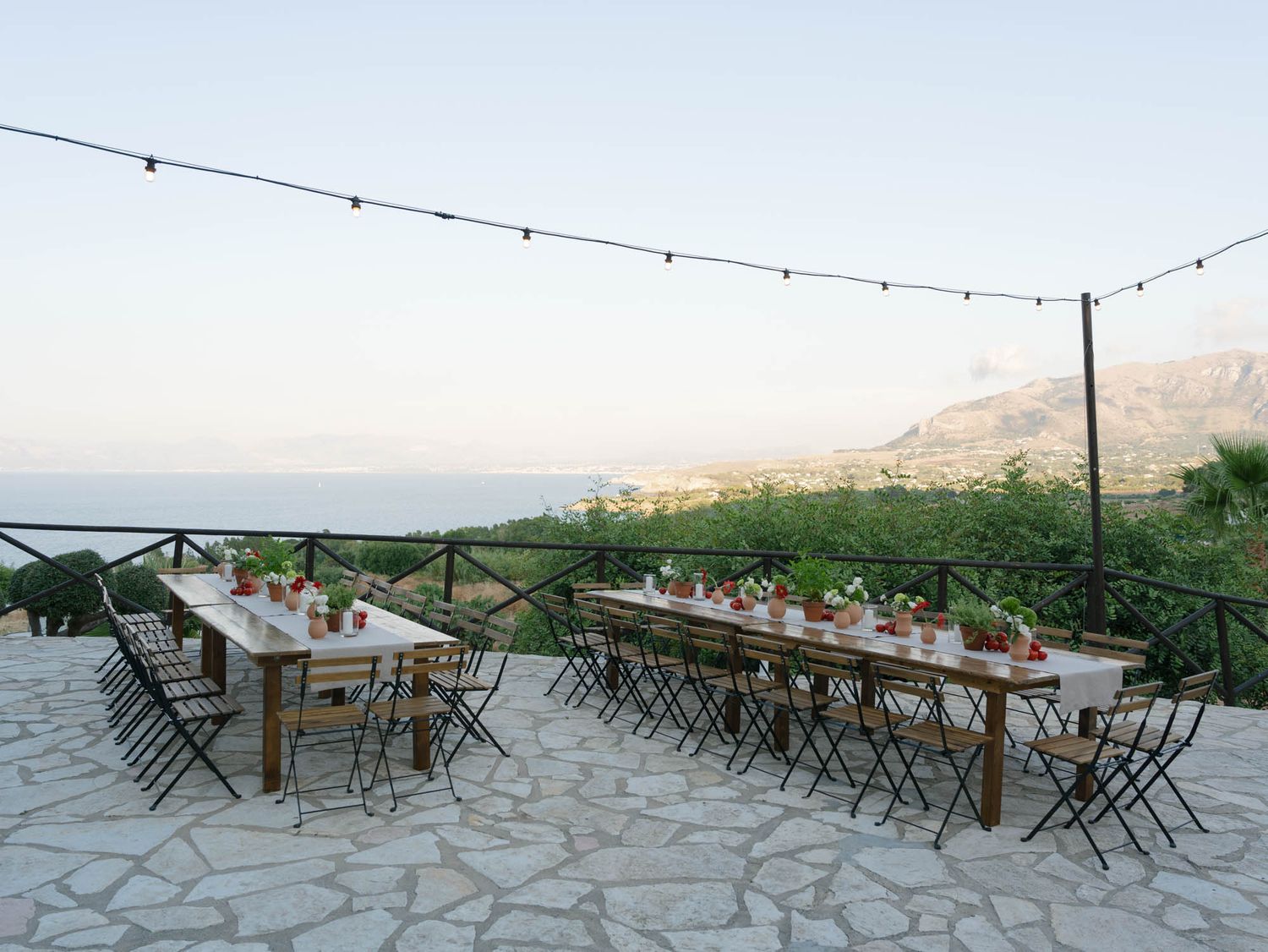 Outdoor dining setup with long tables, string lights, and mountain sea views on a stone terrace. Mulino 1220 in Scopello.