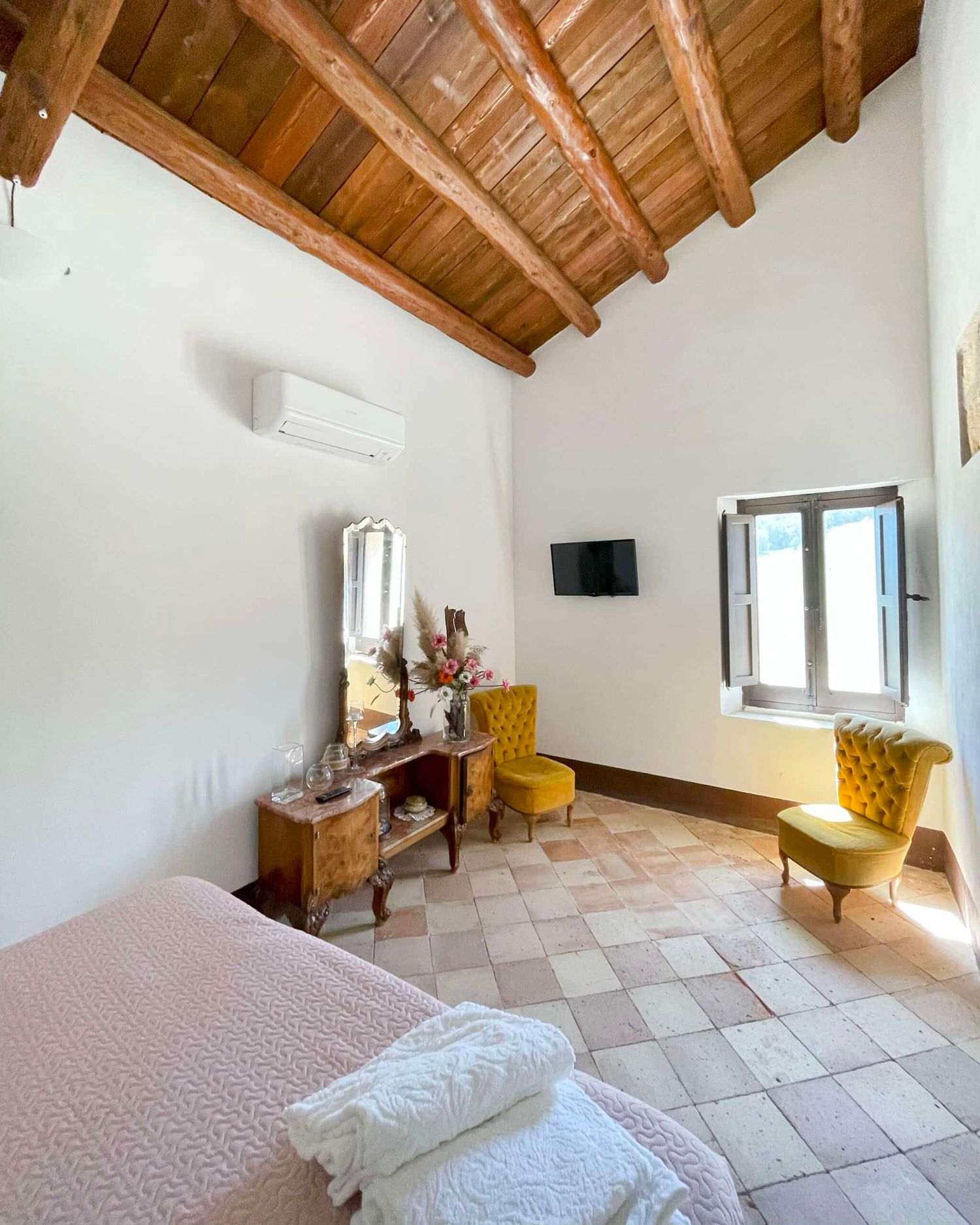 Rustic bedroom with exposed wooden beam ceiling, terracotta tile floor, yellow chairs, and air conditioning unit.
