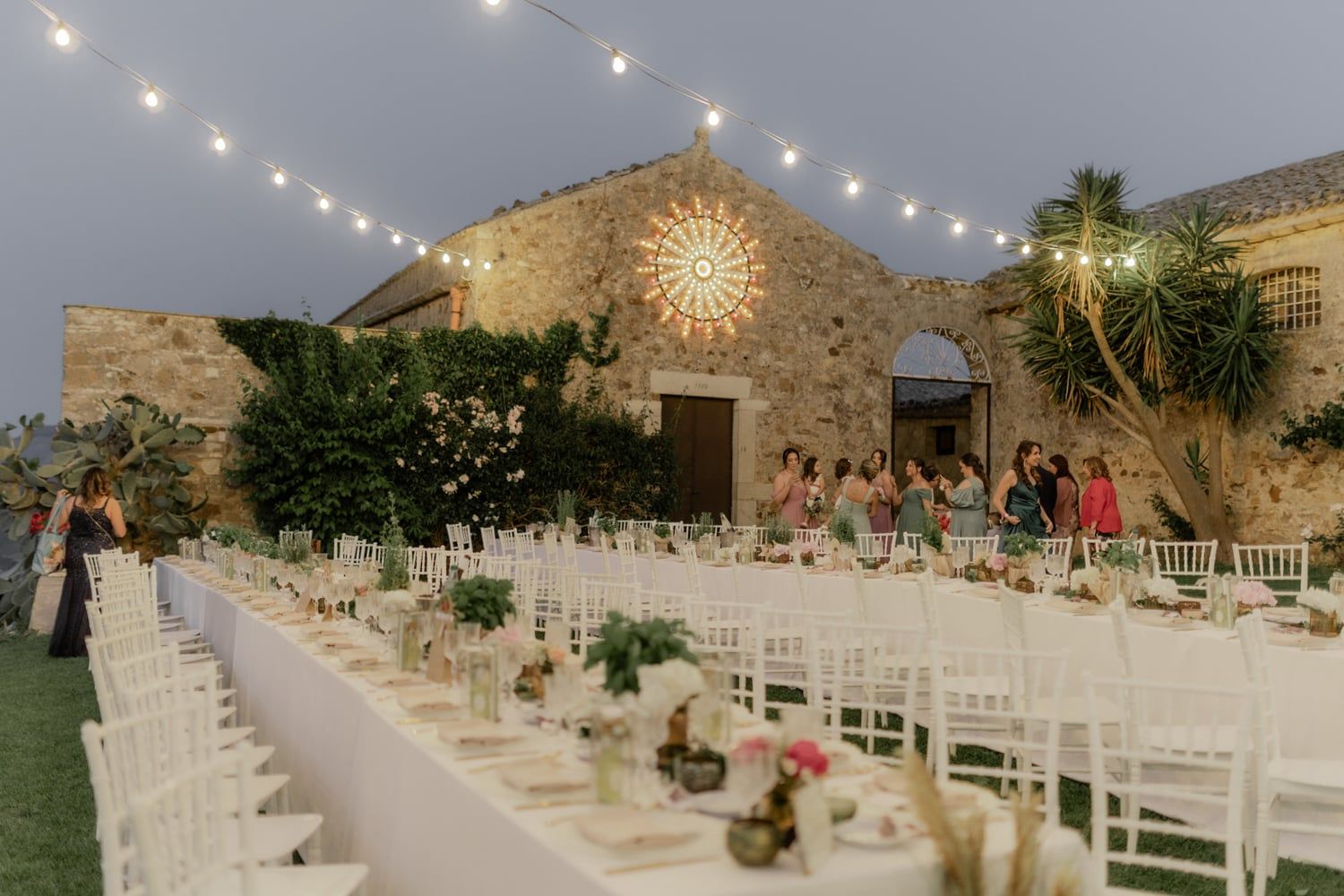 Elegant outdoor wedding reception with string lights, long white tables, and stone building backdrop at dusk.