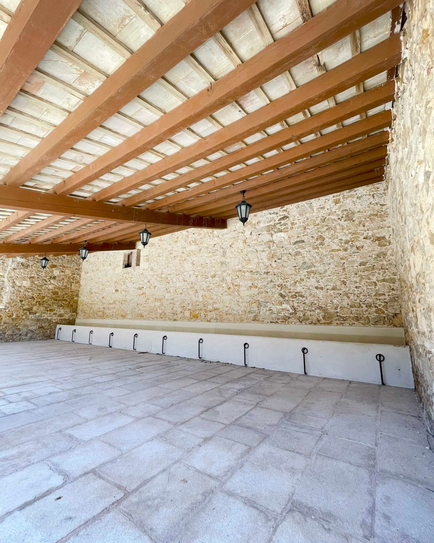 Covered outdoor terrace with wooden pergola, hanging lanterns, and stone wall at a rustic Mediterranean venue.