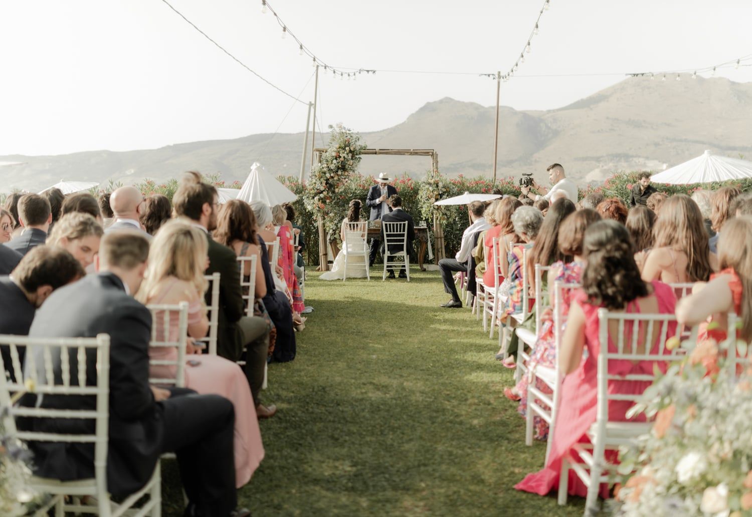 Elegant outdoor wedding ceremony with floral arch, guests in colorful attire seated on white chairs amid mountain backdrop.