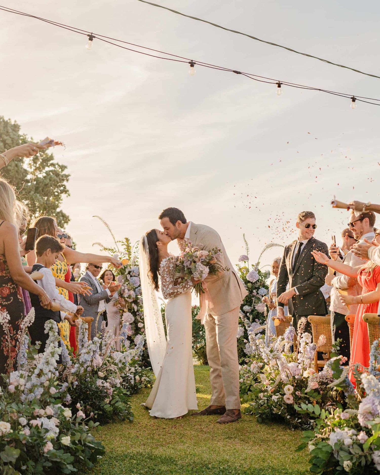 Wedding couple kissing at outdoor ceremony surrounded by white floral arch and cheering guests.
