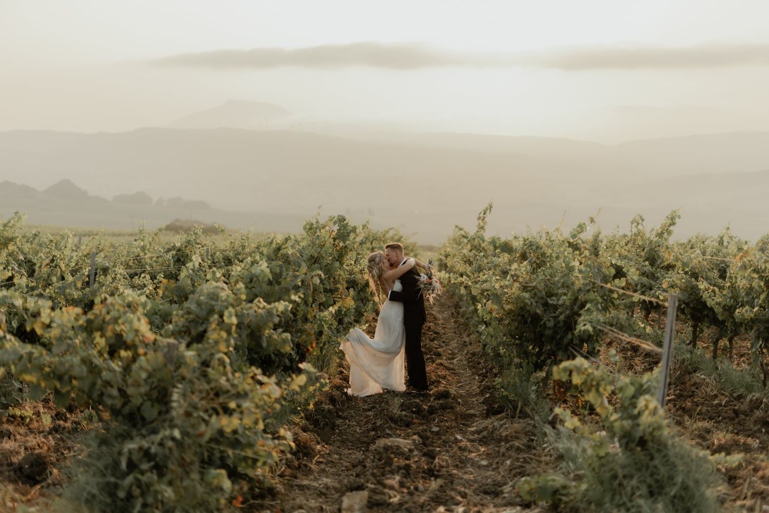 Couple sharing a romantic kiss in a lush vineyard with misty mountains in the background on their wedding day.