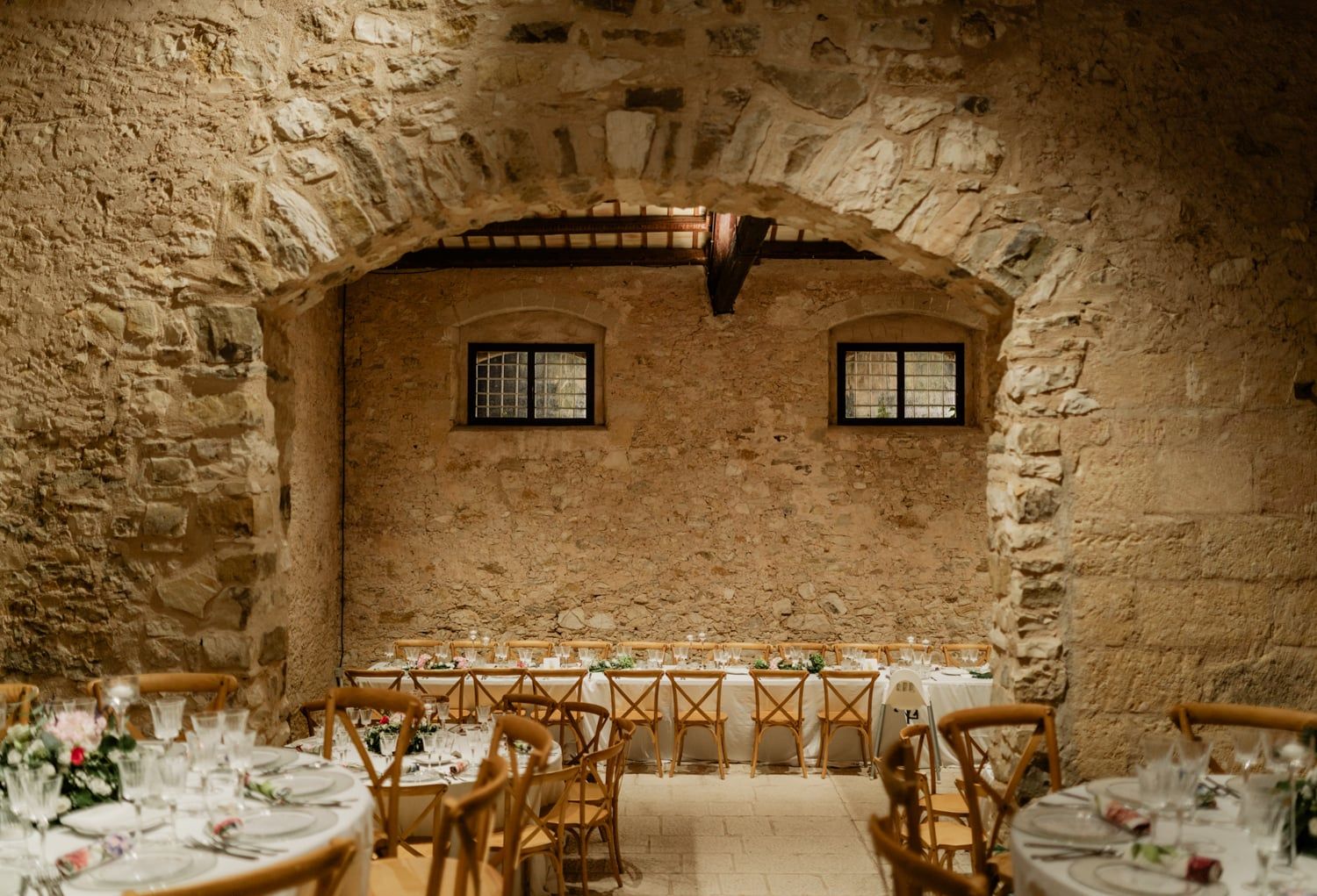 Intimate stone-walled wedding reception hall with arched ceiling, round tables, cross-back chairs, and floral centerpieces.