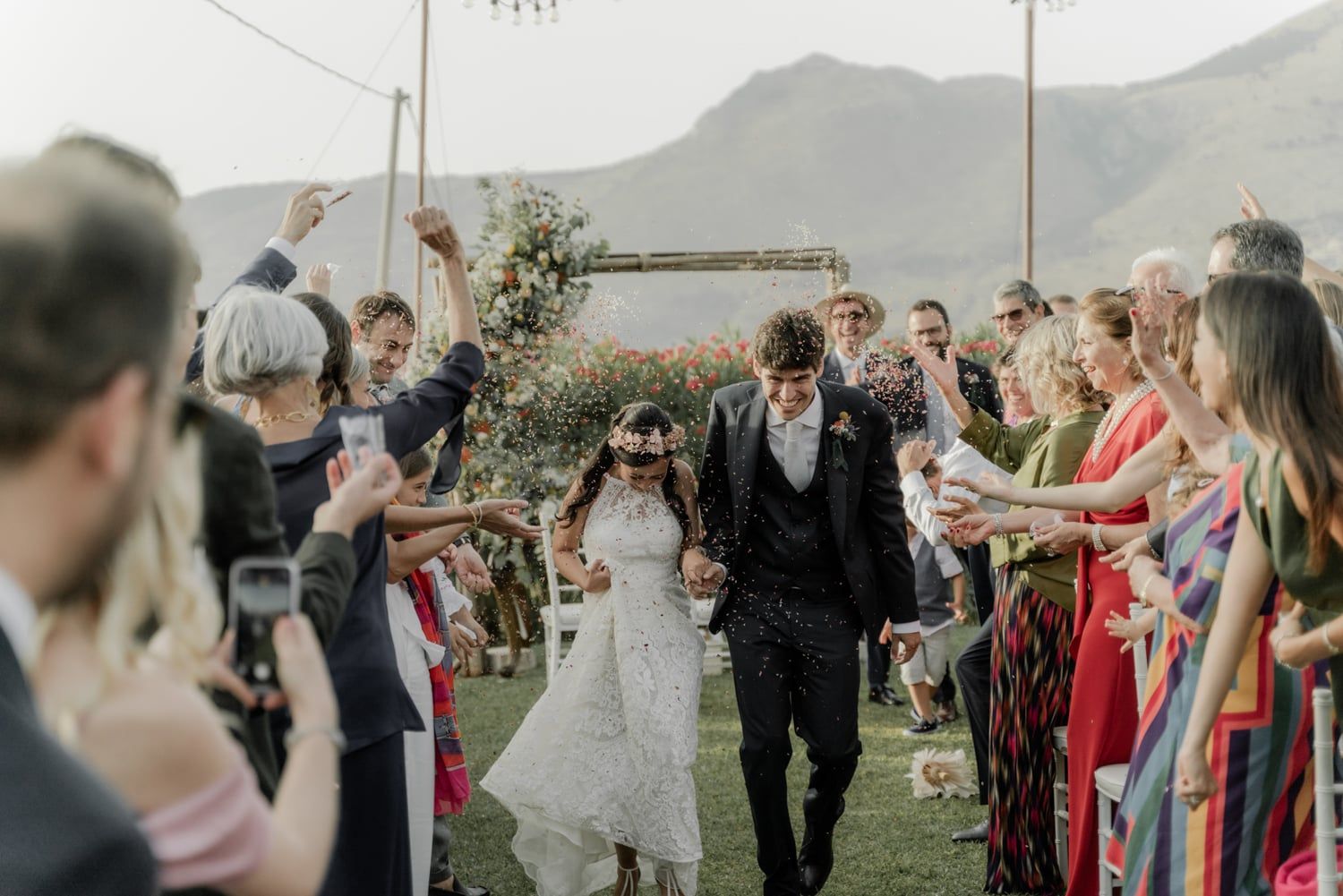 Newlyweds walk through cheering guests tossing confetti after outdoor ceremony with scenic mountain backdrop.