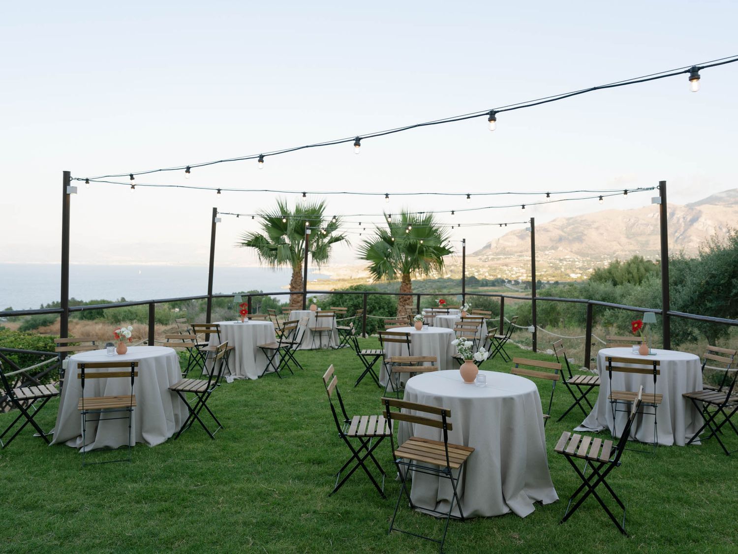Outdoor wedding reception setup with round tables, string lights, and palm trees against a scenic mountain backdrop. Mulino 1220 in Scopello
