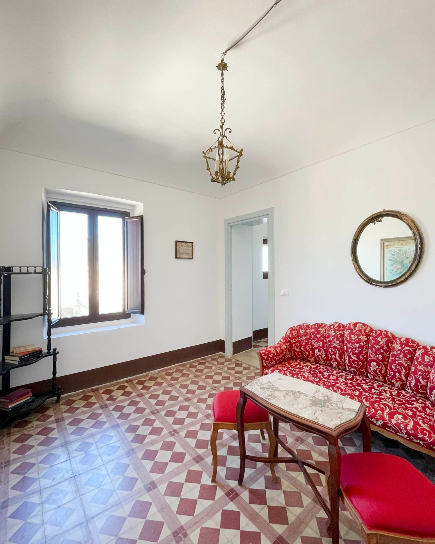 Classic sitting room with red floral sofa, checkered tile floor, brass pendant light, and circular gold mirror.