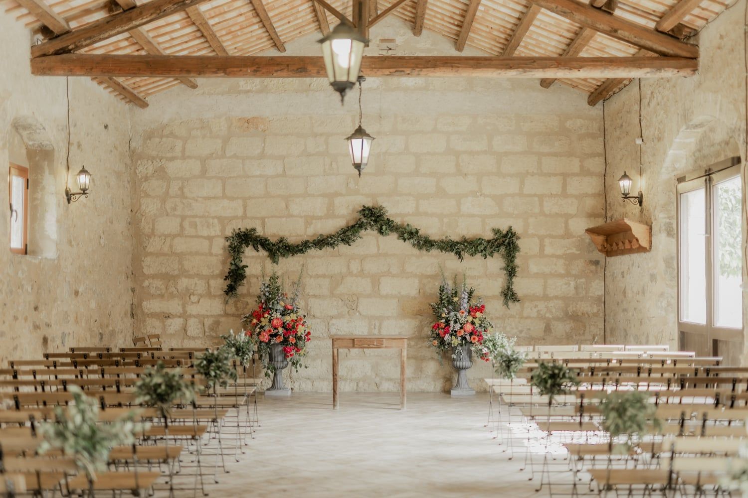 Rustic stone wedding ceremony venue with wooden chairs, floral arch, and hanging lanterns.
