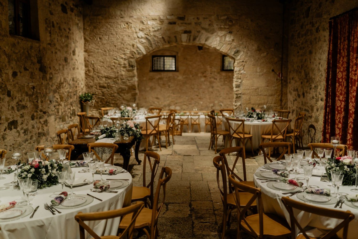 Intimate wedding reception inside a rustic stone-walled venue with round tables, wooden chairs, and candlelit floral centerpieces.