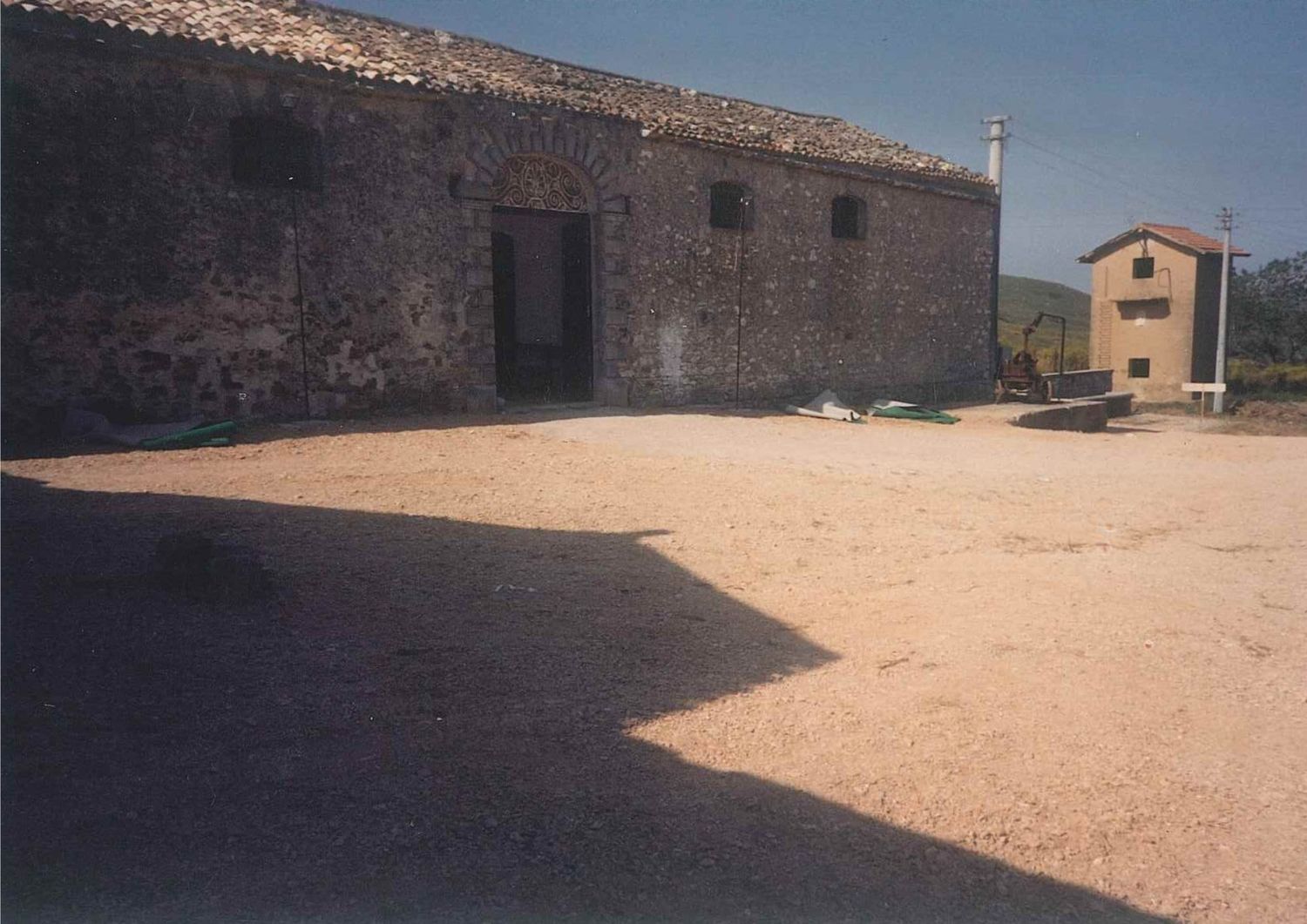 Traditional rustic stone barn with large gravel courtyard under bright sunlight in rural countryside.