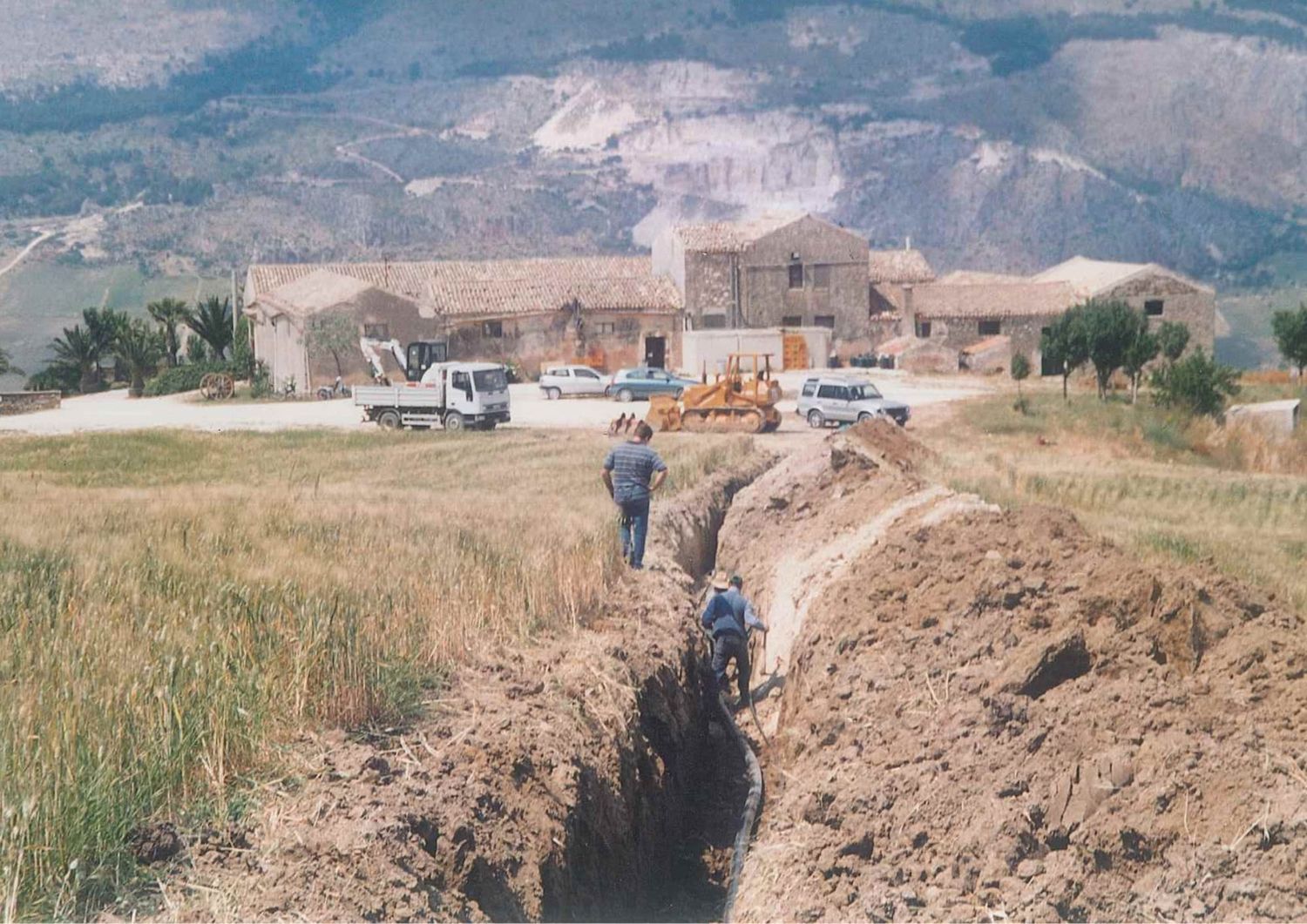 Construction workers digging a trench near a rustic stone estate with mountain backdrop.