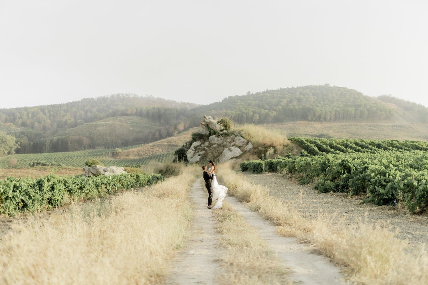Bride and groom share romantic moment on scenic vineyard path with misty hills and lush green vines in background.
