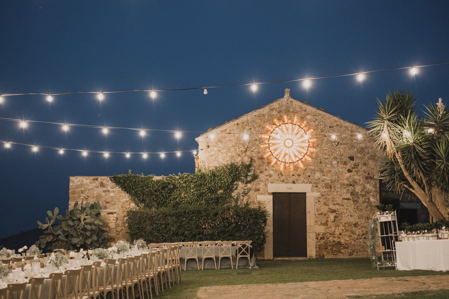 Elegant outdoor wedding reception with string lights illuminating a rustic stone chapel at night.