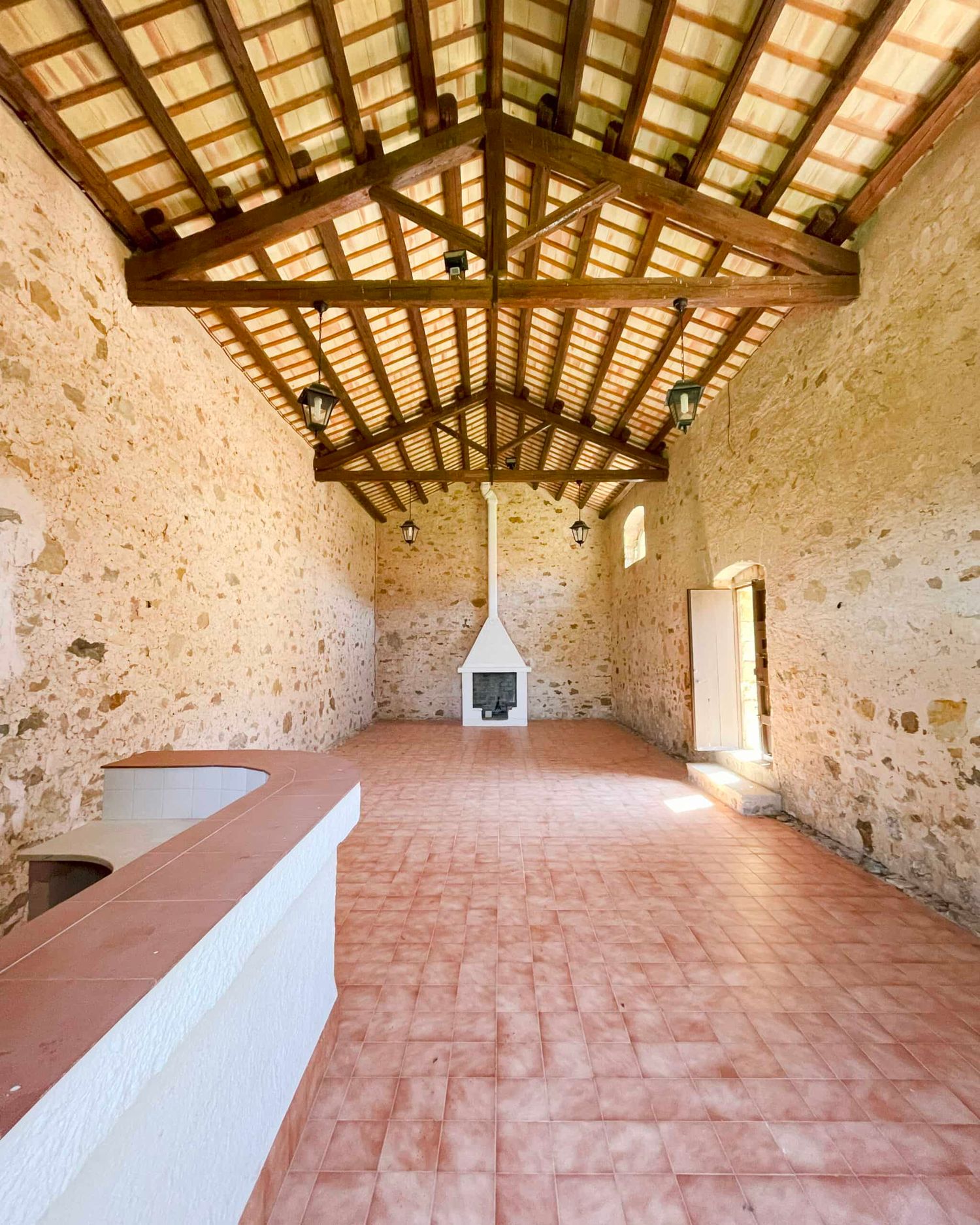 Rustic stone interior with terracotta tile floor, vaulted wooden beam ceiling, and a white fireplace in a renovated farmhouse.