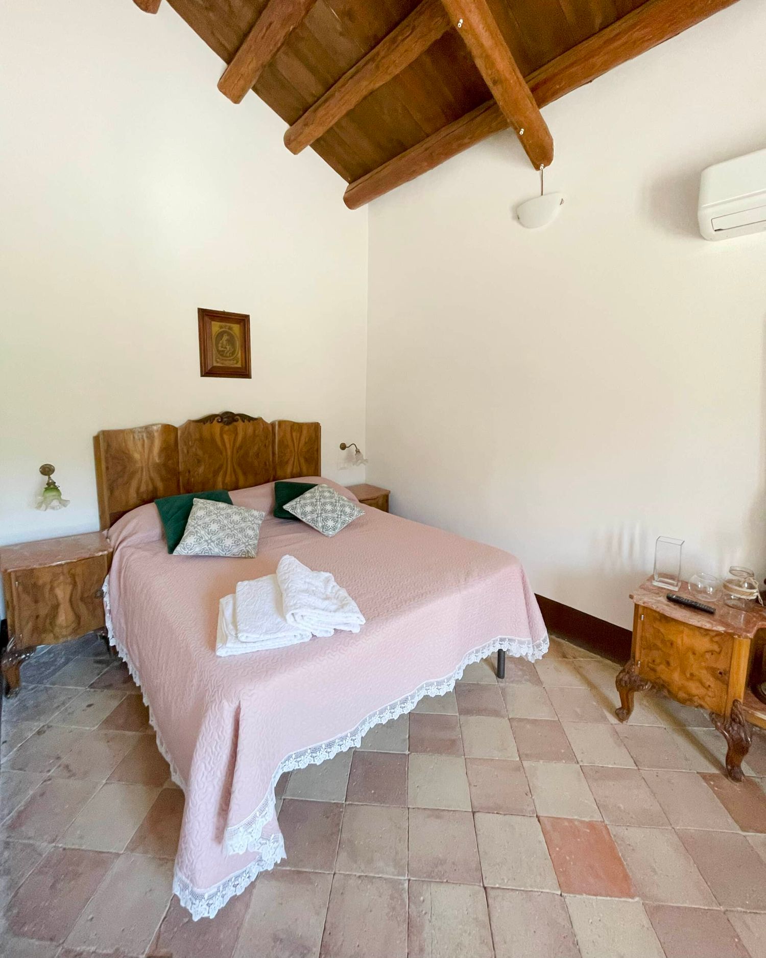 Cozy rustic bedroom with pink bedspread, wooden beam ceiling, antique headboard, and terracotta tile flooring.
